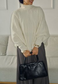 Personne en pull côtelé crème et pantalon gris tenant un sac à main en cuir noir, debout devant un canapé gris clair.