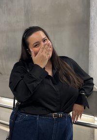 Mujer con cabello largo y oscuro, vistiendo blusa negra y vaqueros azules, sonriendo con la mano parcialmente cubriendo la boca, apoyada en una barandilla de metal.