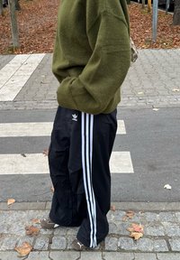 Maglione verde oliva lavorato a maglia, pantaloni cargo neri con tre strisce bianche e scarpe slip-on marroni, in piedi su un marciapiede.