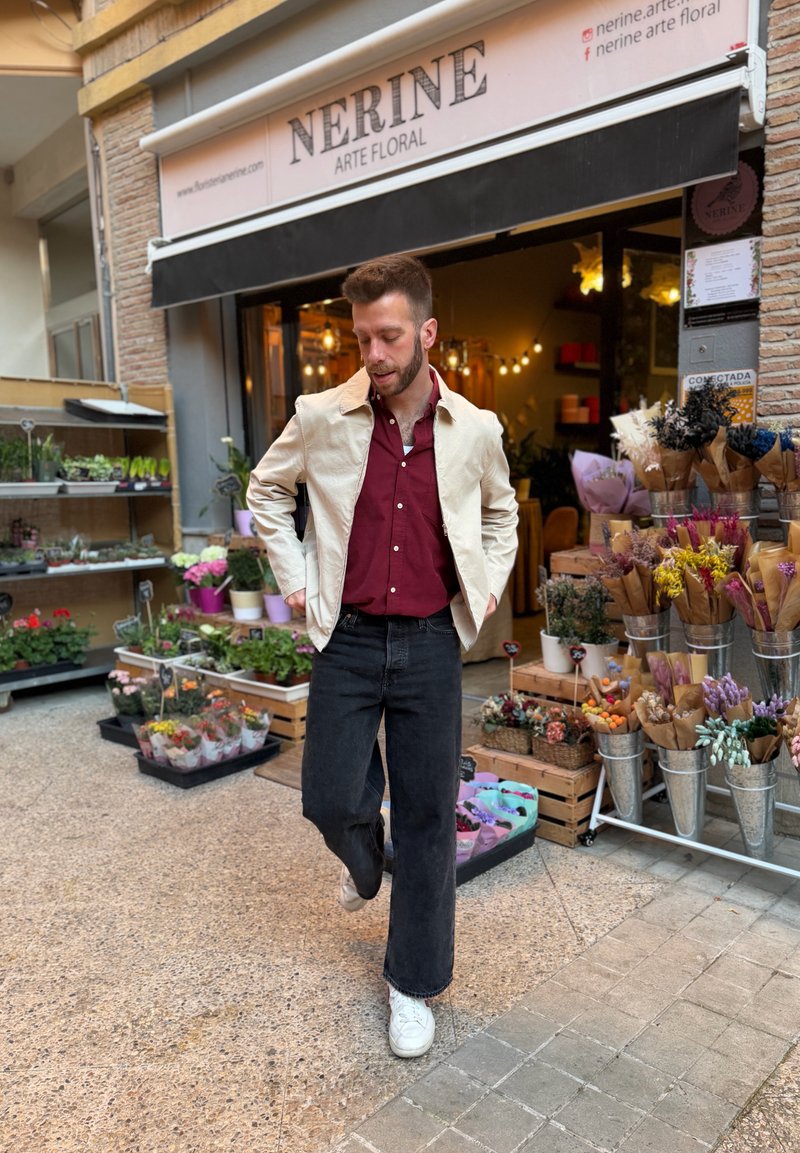 Man in white jacket and red shirt standing outside Nerine floral shop with various potted plants and flower bouquets on display.