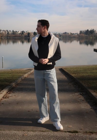 Hombre con un suéter negro y una prenda blanca sobre los hombros de pie en un sendero junto al lago, mirando hacia su izquierda en un día soleado.