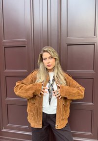 Brun semsket skinnjakke med avslappet passform, med store lommer. Under den, en hvit topp med et leopardmønstret skjerf. Mørke bukser fullfører antrekket.