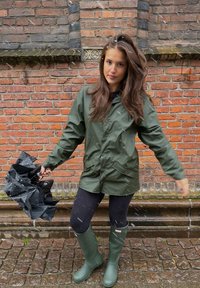 Giacca impermeabile verde con chiusura a bottoni, leggings neri e stivali di gomma verdi, che tiene un ombrello nero chiuso, davanti a un muro di mattoni.