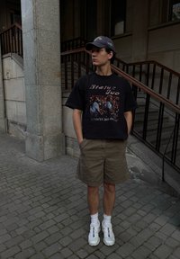 Młody mężczyzna w czarnej koszulce zespołu Status Quo, w khaki szortach, białych trampkach i granatowej czapce, stoi z rękami w kieszeniach na paved ulicy.