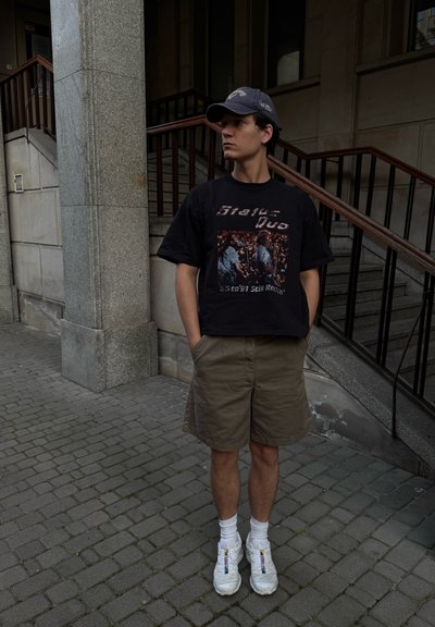 Camiseta gráfica negra con estampado retro de una banda, pantalones cortos color caqui, calcetines blancos y zapatillas blancas voluminosas. Entorno urbano con elementos de piedra y madera.