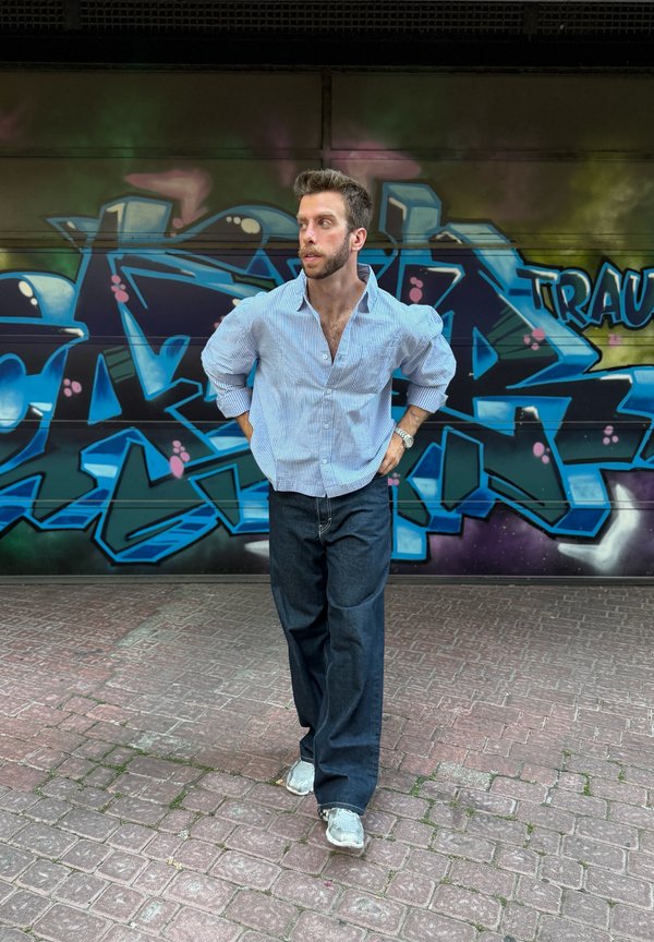 Man met baard en kort haar, gekleed in een gestreept shirt en donkere spijkerbroek, staat op een betegelde straat voor een blauwe graffiti muur.