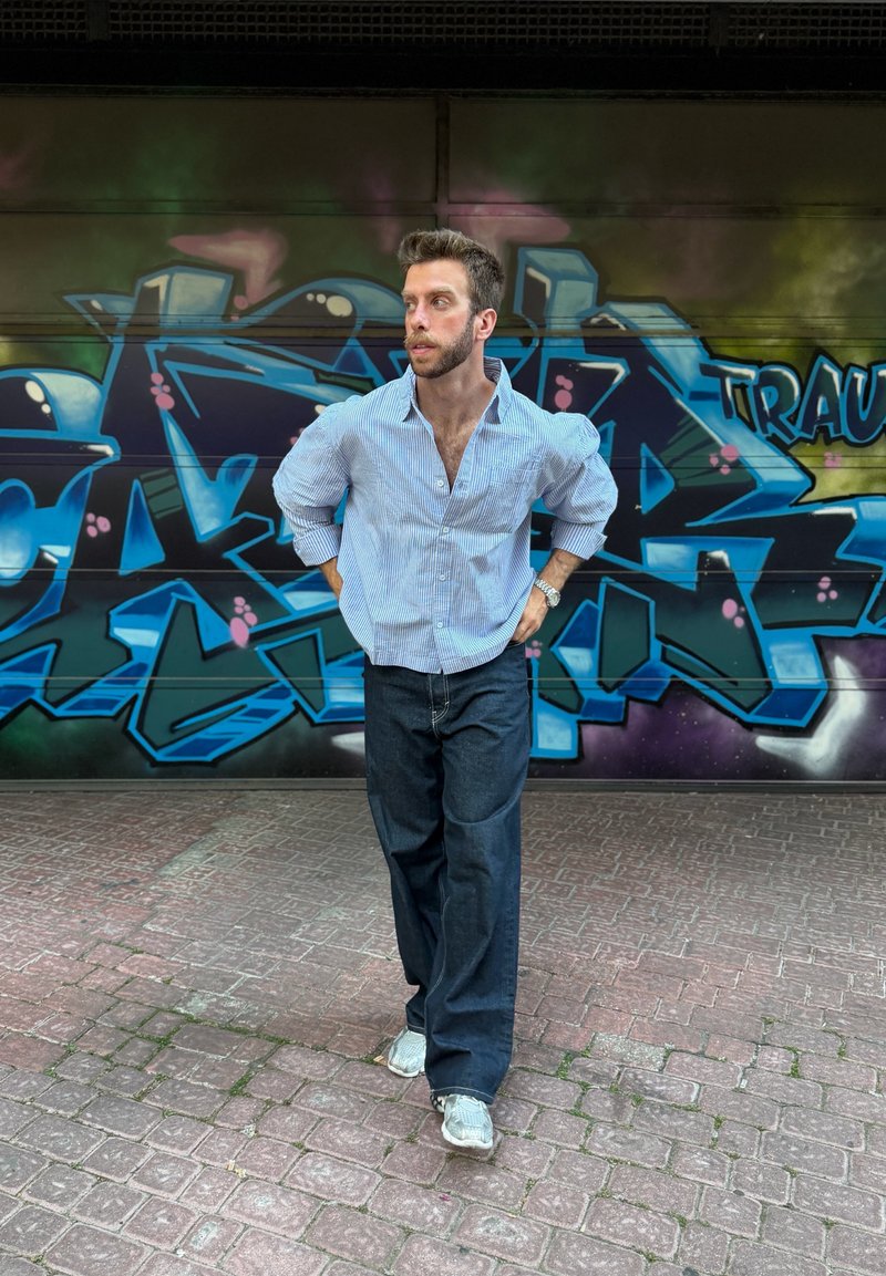 Homme avec barbe et cheveux courts portant une chemise rayée et un jean foncé, debout dans une rue carrelée devant un mur de graffiti bleu.