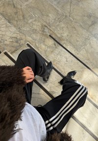 Personne assise sur des escaliers en marbre, portant un pantalon noir avec des bandes blanches, des chaussures noires pointues, une chemise blanche et une veste sombre en fourrure.