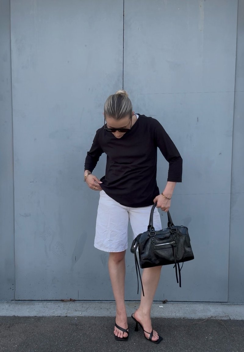 Frau trägt schwarzen Langarmtop, weiße Shorts, schwarze High-Heel-Sandalen, hält schwarze Handtasche, blickt nach unten vor grauer Wand.