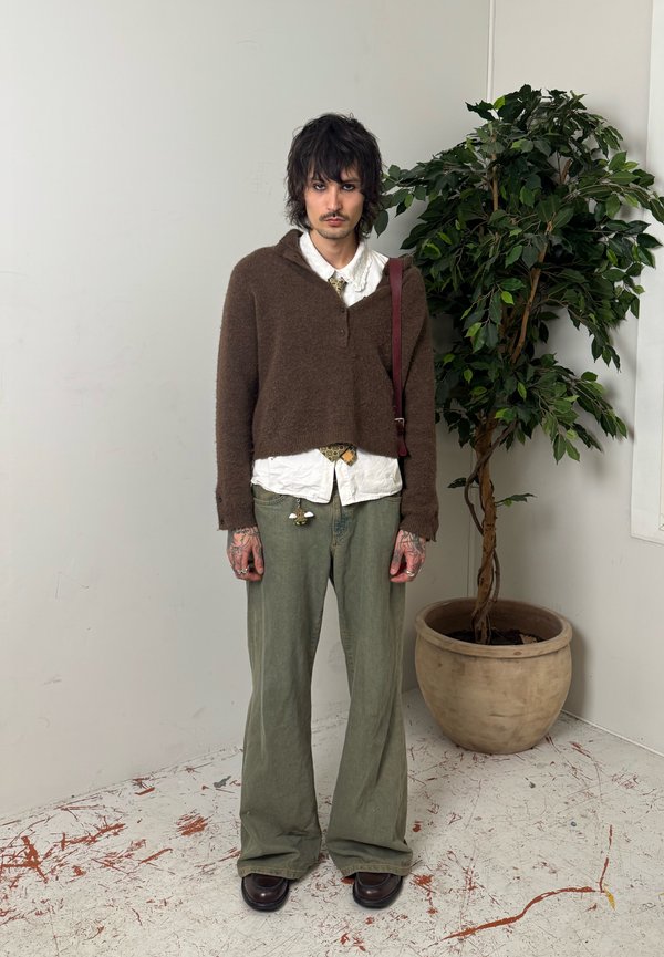 Ung person med ujævnt mørkt hår iført brun sweater, hvid skjorte, løse grønne bukser, stående ved siden af en potteplante med blade i et minimalistisk rum.