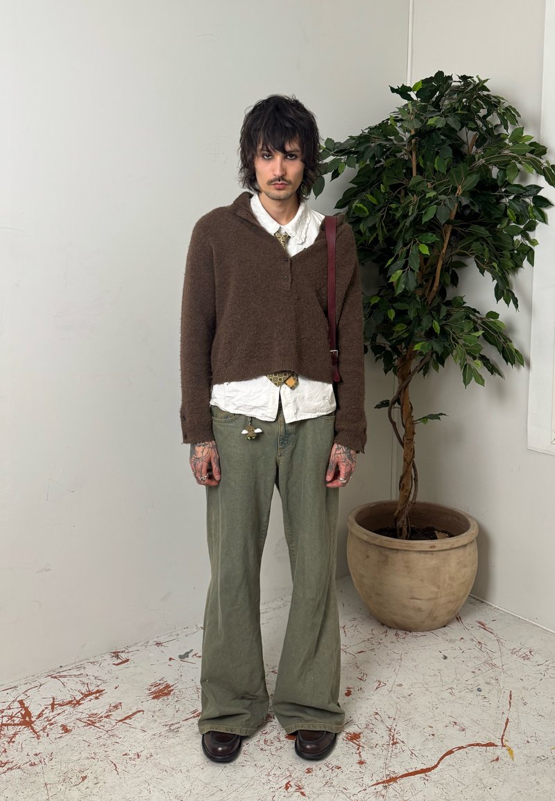 Noor inimene kareda tumeda juustega, kes kannab pruunat kampsunit, valget särki ja lahtisi rohelisi pükse, seisab minimalistikus ruumis potitaime kõrval.