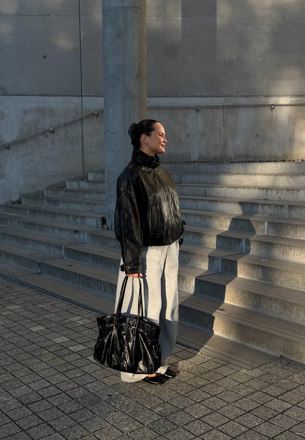 Femme en veste en cuir noire et pantalon gris tenant un grand sac noir, debout près de marches en béton au soleil avec une ombre sur les escaliers.