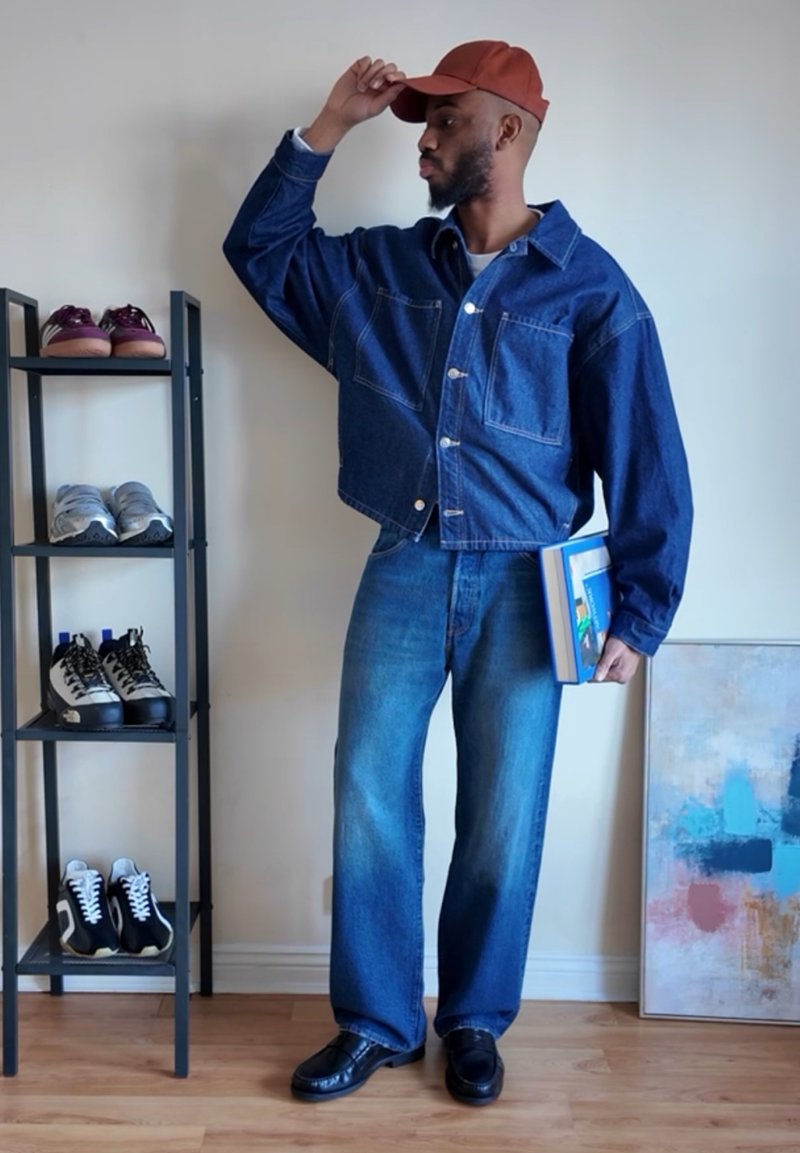 Homme en veste en jean et jeans tient des livres dans une main et ajuste une casquette rouge de l'autre, debout à côté d'un porte-chaussures et d'une peinture abstraite.