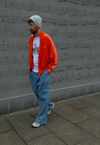 Orange fuzzy Adidas jacket with white stripes, graphic t-shirt, blue baggy jeans, and grey beanie, walking on a stone pavement.