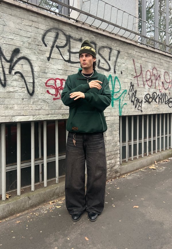 Sweatshirt verde com fecho, com um acabamento texturizado, combinado com calças de denim pretas largas e sapatos pretos. Um porta-chaves pendurado nas calças.