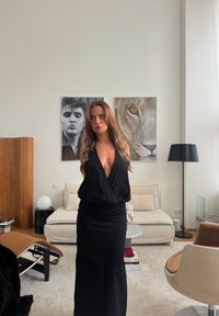 Zwarte jurk met een diepe V-hals, slanke pasvorm en een geplooide taille. Zachte stof contrasteert met de gladde meubels en kunstwerken op de achtergrond.