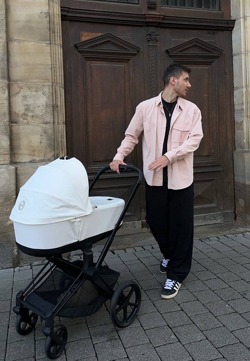 Balts bērnu ratiņš ar noapaļotu kapuci, melnu pamatni un lieliem riteņiem. Vīrietis rozā krekliņā un melnās biksēs stāv blakus pie koka durvīm.