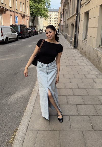 Falda maxi de mezclilla con una abertura frontal y dobladillo deshilachado, combinada con una blusa negra ajustada y zapatos negros. Un bolso negro está colgado de un hombro.