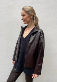 Brązowa skórzana kurtka z wysokim kołnierzem i zapięciem na zamek. Charakteryzuje się gładką fakturą i lekko skróconym krojem, w zestawieniu z czarną bluzką.