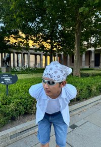 Individuo che indossa un bandana bianca con motivi neri, occhiali da sole, una t-shirt bianca e una camicia a righe blu, abbinati a pantaloni corti di denim.