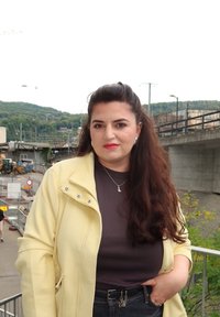 Žena s dugom tamnom kosom nosi žutu jaknu i tamnu majicu, stoji na otvorenom blizu gradilišta i zelenih brda.