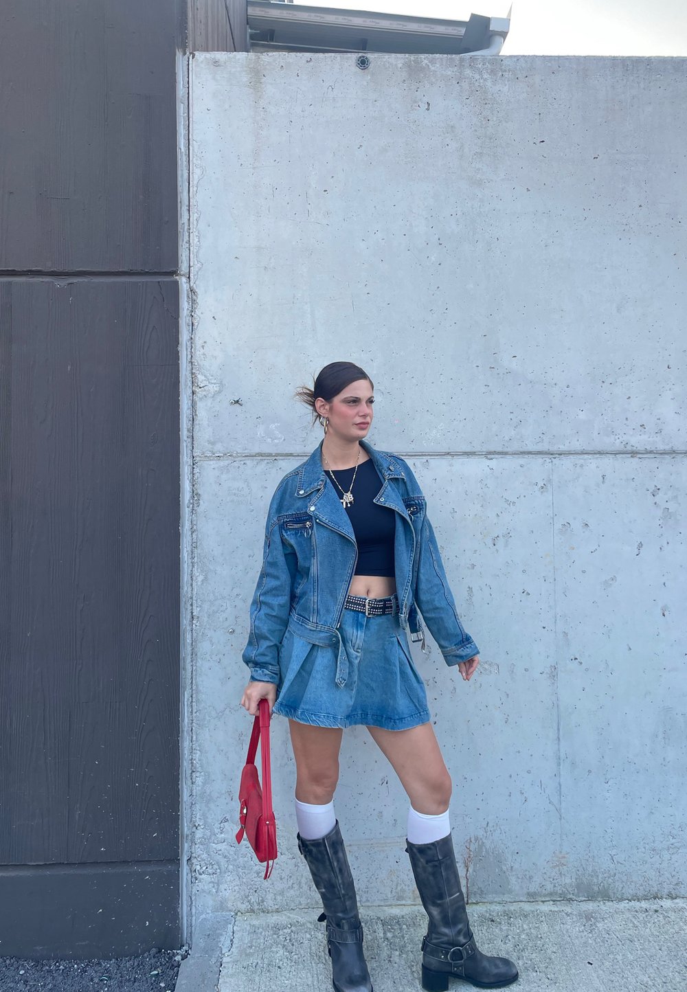 Jeans jakna z zippers, črni kratki top, jeans krilo, rdeča torbica, črne škornje do kolen in bele kolenske nogavice ob sivi steni.