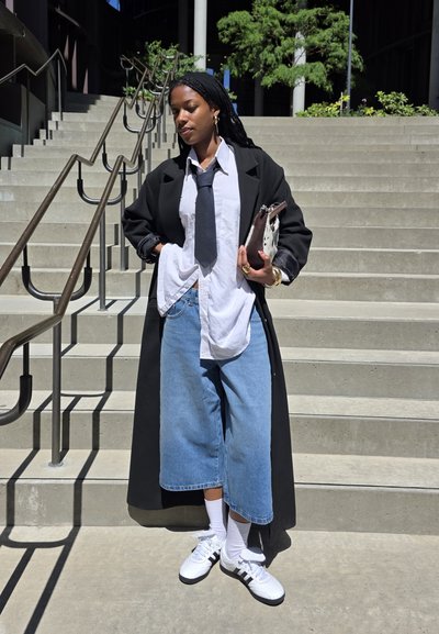 Abrigo largo negro, camisa blanca, corbata oscura, culottes de denim de gran tamaño y zapatillas blancas con rayas negras. Sosteniendo un bolso de mano, de pie en las escaleras.