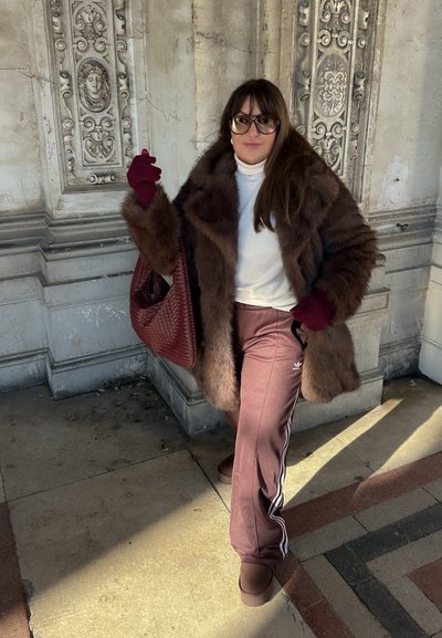 Abrigo de piel sintética marrón, suéter blanco de cuello alto, bolso de cuero sintético burdeos, guantes burdeos, pantalones de chándal marrones de Adidas con rayas blancas y botas marrones.