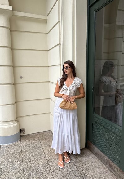 Mujer con blusa blanca de lunares y falda larga blanca sostiene un clutch de mimbre, lleva gafas de sol y sandalias, apoyada contra una pared crema junto a una puerta verde.