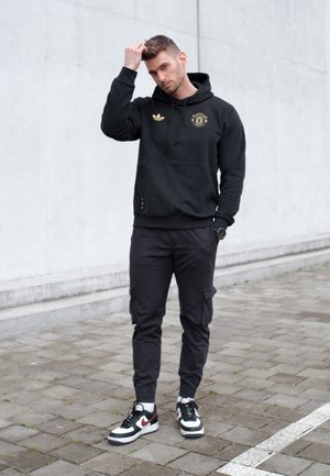 Joven con sudadera negra con capucha y pantalones cargo que se ajusta el cabello, de pie sobre un pavimento gris frente a una pared lisa de color claro.