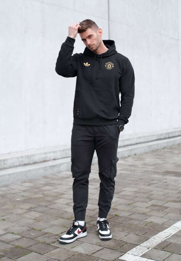 Jaunas vyras juodu gobtuvu ir krovinių kelnėmis susitvarko plaukus, stovėdamas ant pilkos dangos priešais paprastą šviesią sieną.