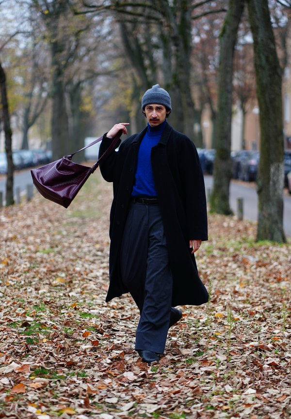 Man walking on leaf-covered path between rows of trees, wearing a gray beanie, blue turtleneck, black coat, and carrying a large maroon bag.