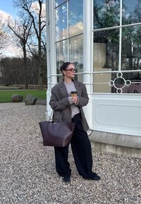 Bruine wollen blazer over een grijze trui, zwarte wijde broek en een grote leren tote bag in bruin. Houdt een koffie-to-go-beker vast.
