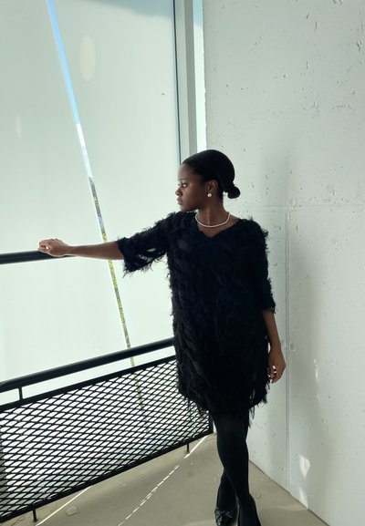 Vestido negro de plumas con un corte holgado, mangas cortas y escote en V. La modelo lleva medias negras y un collar de perlas, de pie cerca de una barandilla.