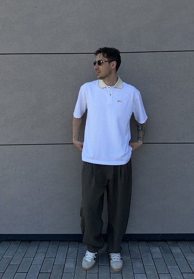 Hombre con gafas de sol, camiseta polo blanca, pantalones oscuros y zapatillas blancas, de pie contra una pared de azulejos grises con las manos en los bolsillos.
