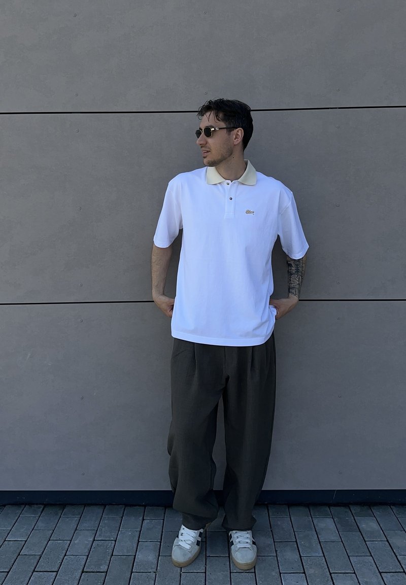 Homme portant des lunettes de soleil, un polo blanc, un pantalon foncé et des baskets blanches, debout contre un mur carrelé gris, les mains dans les poches.