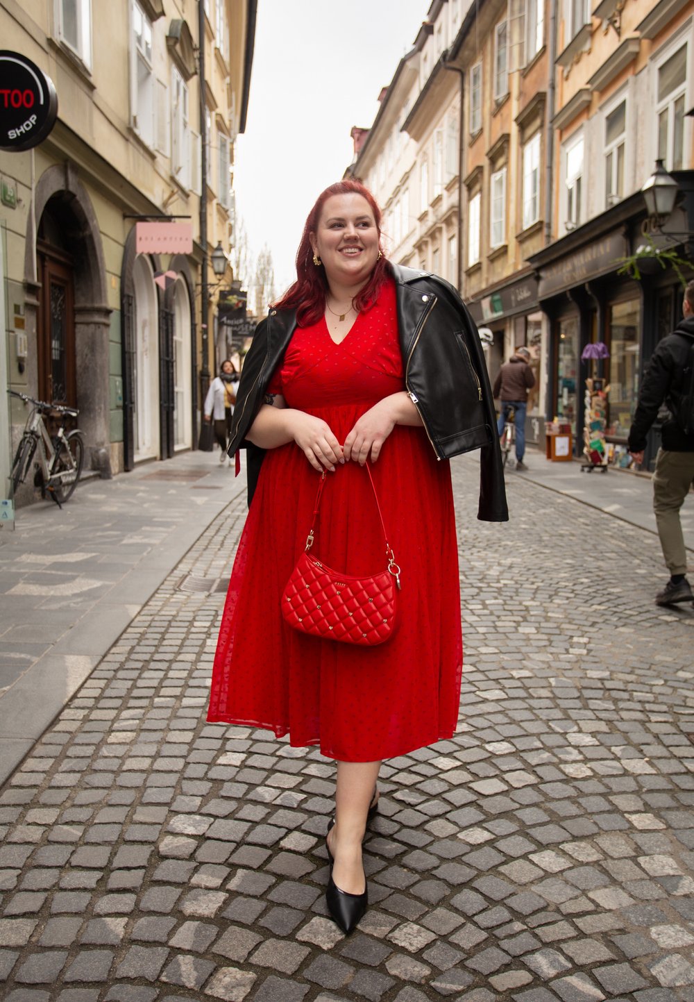 Vestido rojo con lunares, escote en V, falda fluida, combinado con una chaqueta de cuero negra. Sostenido un bolso acolchado rojo. Fondo de calle adoquinada.