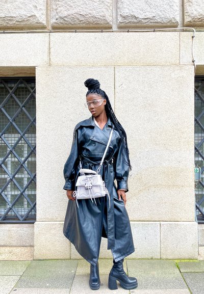 Abrigo de trench negro de cuero con cuello sobredimensionado, cinturilla ceñida y mangas largas; combinado con botas de plataforma gruesas y un bolso gris claro.