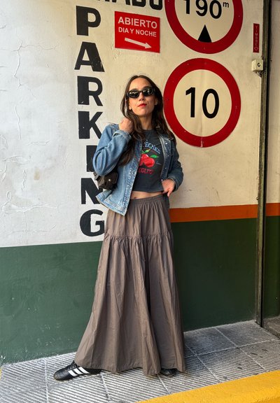 Chaqueta de denim, camiseta gráfica con cerezas y una falda larga marrón con volantes. Zapatos negros con rayas blancas. Fondo de hormigón con señales de aparcamiento.