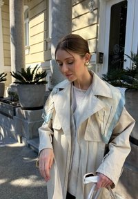 Trench coat crema con accenti blu e linee nere. Presenta un ampio colletto e tasche grandi, abbinato a una blusa leggera e una borsa bianca.