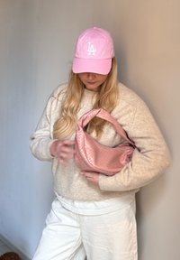 Rosa geflochtene Handtasche mit kurvenförmigem Design und weicher Textur. Die Person trägt einen hellbeigen Pullover und weiße Hose, sowie eine pinke Mütze.