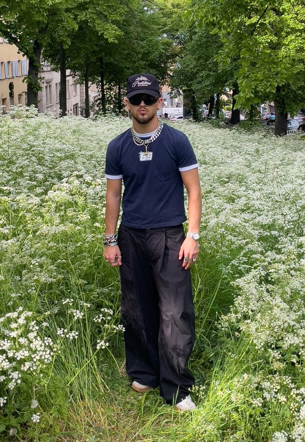 Homme portant une casquette noire, des lunettes de soleil, une chemise bleu foncé et un pantalon noir se tenant dans un champ vert avec des fleurs sauvages blanches et des arbres à proximité.