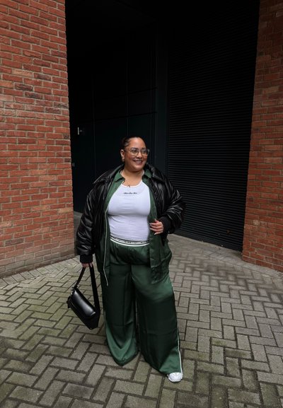 Mujer con pantalones de satén verde, camiseta blanca, chaqueta de cuero negra, llevando un bolso negro, de pie entre paredes de ladrillo en un camino pavimentado.