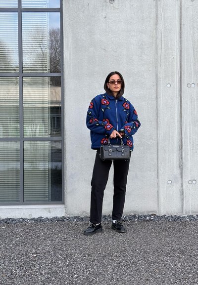 Chaqueta de forro polar azul con bordado floral, pantalones negros, bolso negro y gafas de sol. Textura suave, cierre de cremallera y corte relajado.