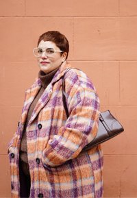 Άτομο με κοντά μαλλιά και μεγάλα γυαλιά, φοράει παλτό καρό σε πορτοκαλί και μοβ αποχρώσεις και κρατάει μία σκούρα καφέ τσάντα, μπροστά σε ένα ροδακινί τοίχο.