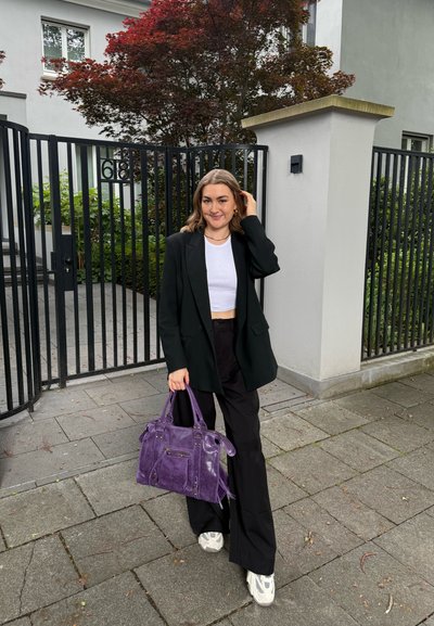 Blazer negro sobre un top corto blanco, pantalones anchos negros, zapatillas grises y un bolso de piel morado con bolsillos laterales, de pie al aire libre.
