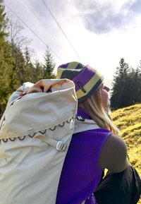 Világos bézs hátizsák, megkötős tetejével és díszes fűzésekkel, lila fleece mellény felett viselve, színes kötött sapkával párosítva.
