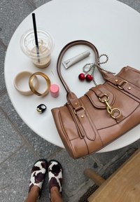 Bolsa de mão de couro castanho com acabamento em dourado, alça decorativa e fecho. Os acessórios incluem pulseiras, um bálsamo labial rosa e uma bebida com palhinha.