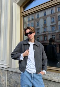 Grau gerippte Jacke über einem weißen Hemd, kombiniert mit hellblauen Jeans. Er trägt schwarze Sonnenbrillen und steht nahe einem Fenster mit Reflexionen.