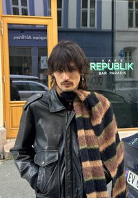 Veste en cuir noire avec fermetures éclair et poches, superposée avec une écharpe rayée et duveteuse en marron, noir et vert. Arrière-plan urbain avec un panneau de café.